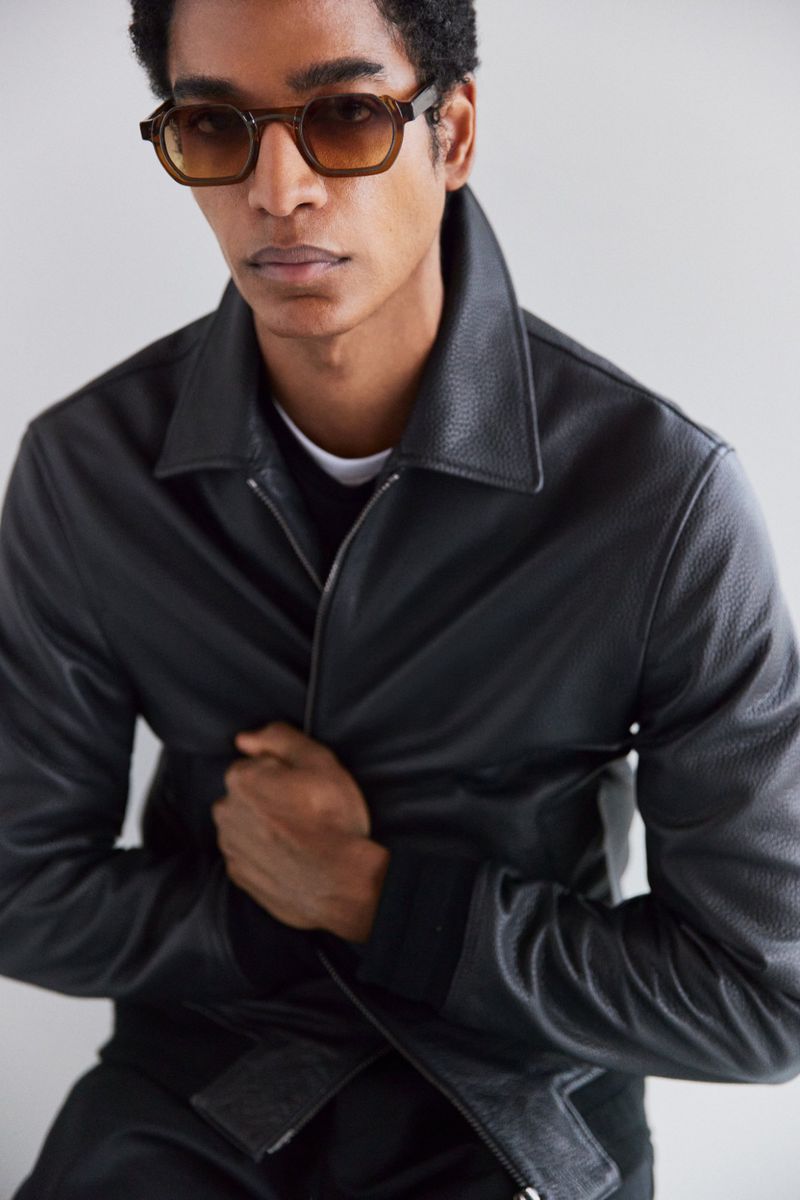 Portrait of an adult Black male wearing sunglasses and a textured black jacket against a neutral background in a fashion photography studio.