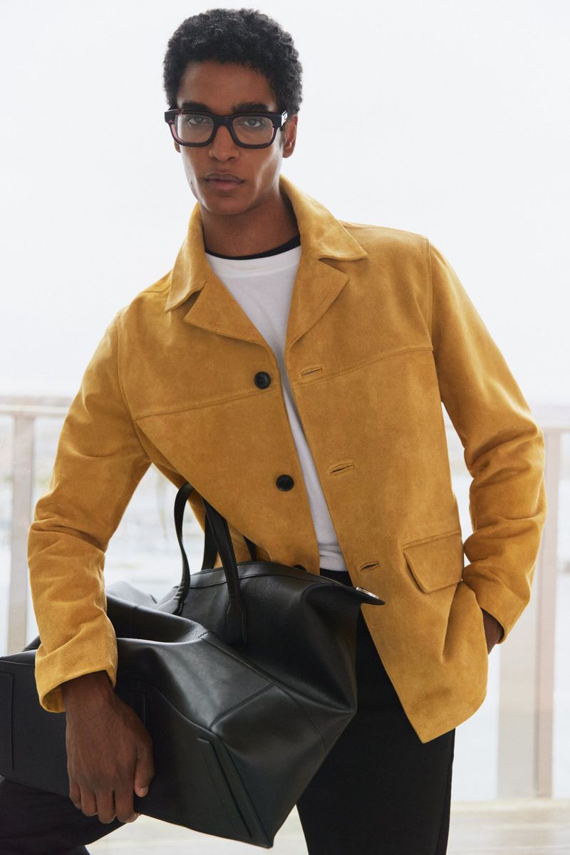 A male model poses indoors wearing a tan jacket and black trousers while holding a black leather duffel bag. He wears glasses.