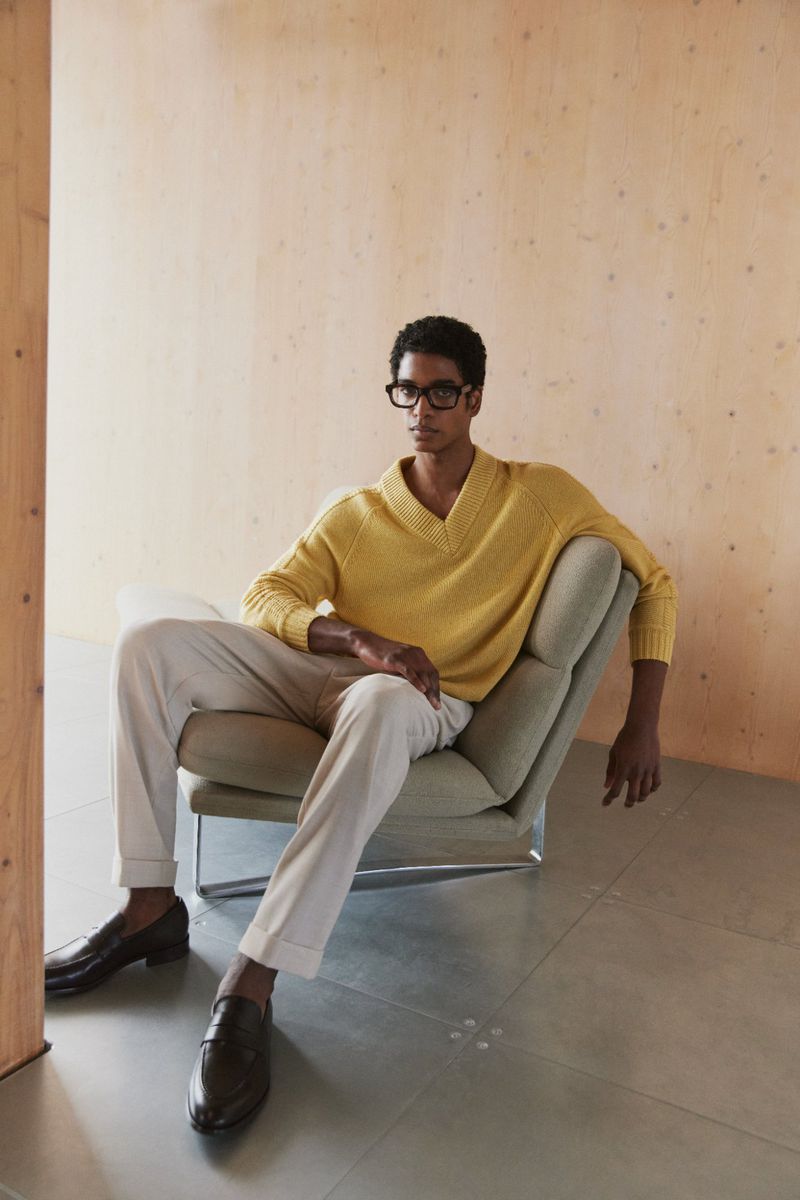 Dark-haired male model in yellow knit sweater, beige slacks, and black loafers sits in a chair against a wood paneled wall.