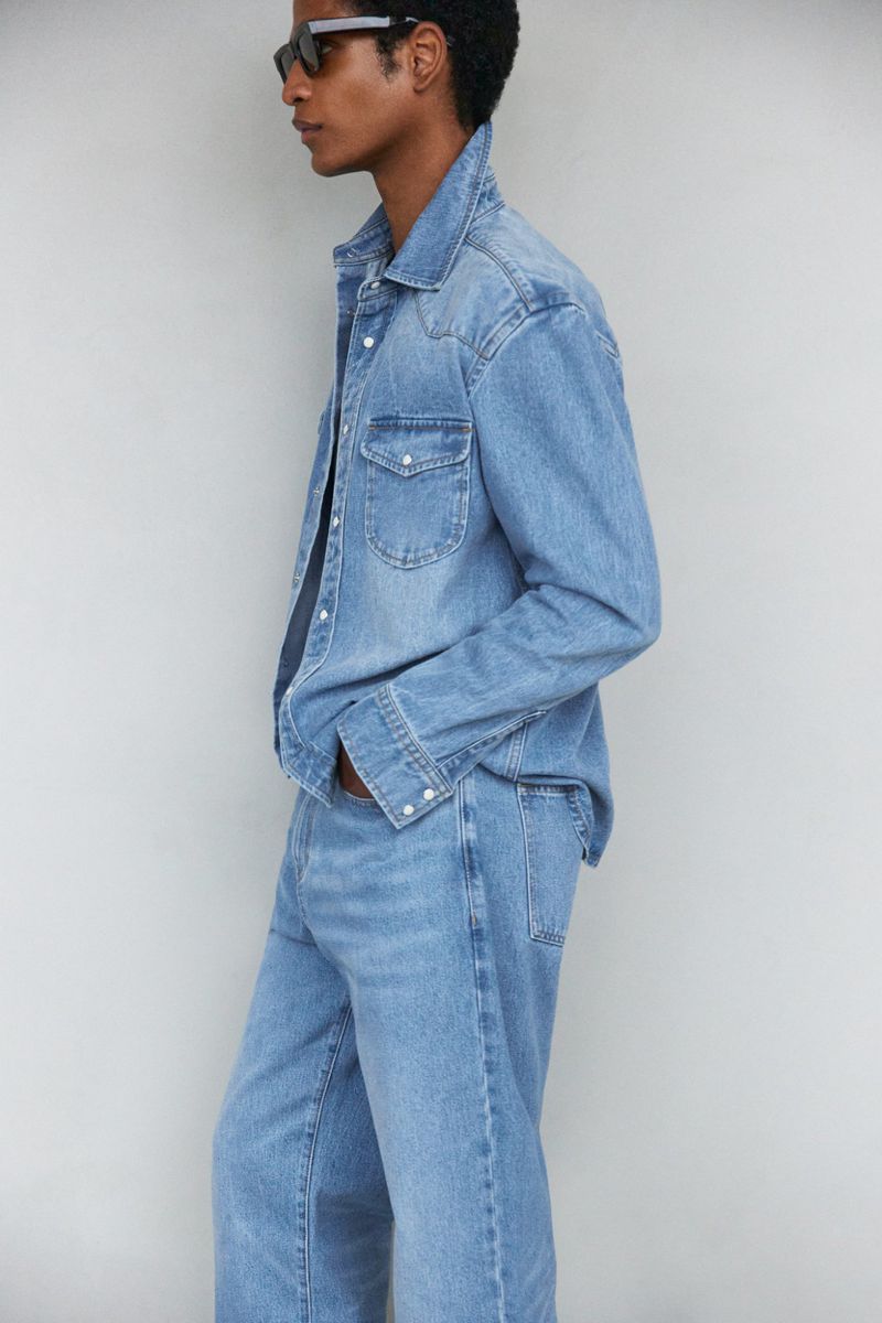 Profile shot of a man wearing a light blue denim shirt, jeans, and black sunglasses, posing against a neutral background.