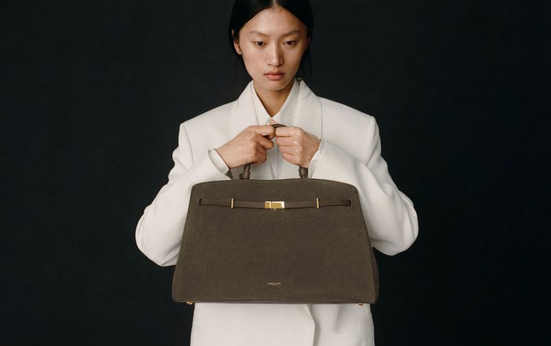 Studio shot of a stylish woman presenting a structured brown leather handbag with gold hardware against a dark background.