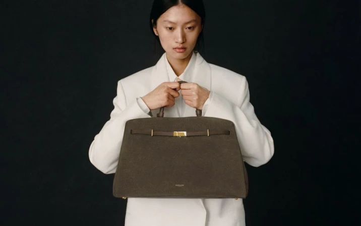 Studio shot of a stylish woman presenting a structured brown leather handbag with gold hardware against a dark background.