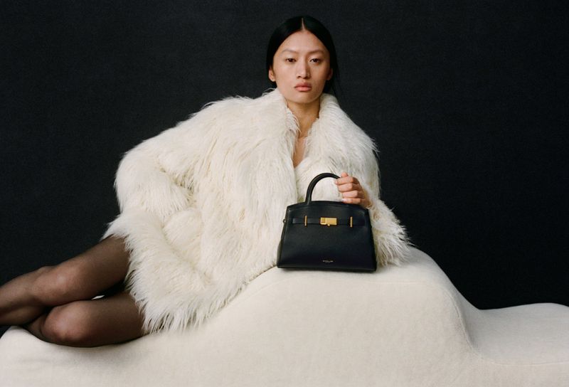 Asian woman poses indoors, wearing a fur coat and holding a black leather handbag against a dark background in a studio shot.