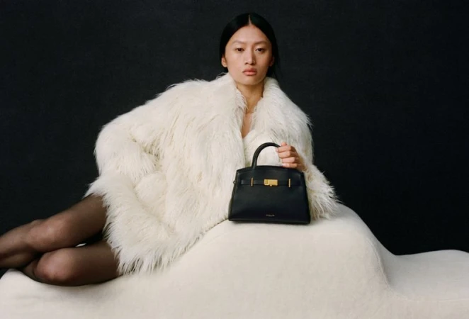 Asian woman poses indoors, wearing a fur coat and holding a black leather handbag against a dark background in a studio shot.