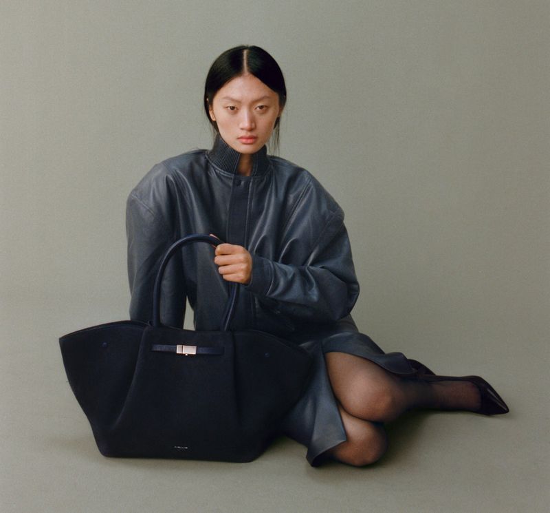 Asian woman sits with black handbag in a studio, wearing a blue leather jacket and black tights, with high heel shoes.