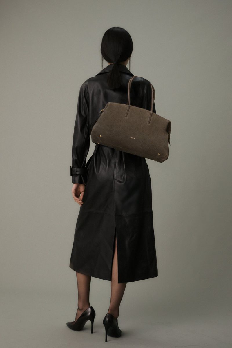 Back view of adult female wearing black leather trench coat, heels, and beret with gray leather handbag in a studio setting.