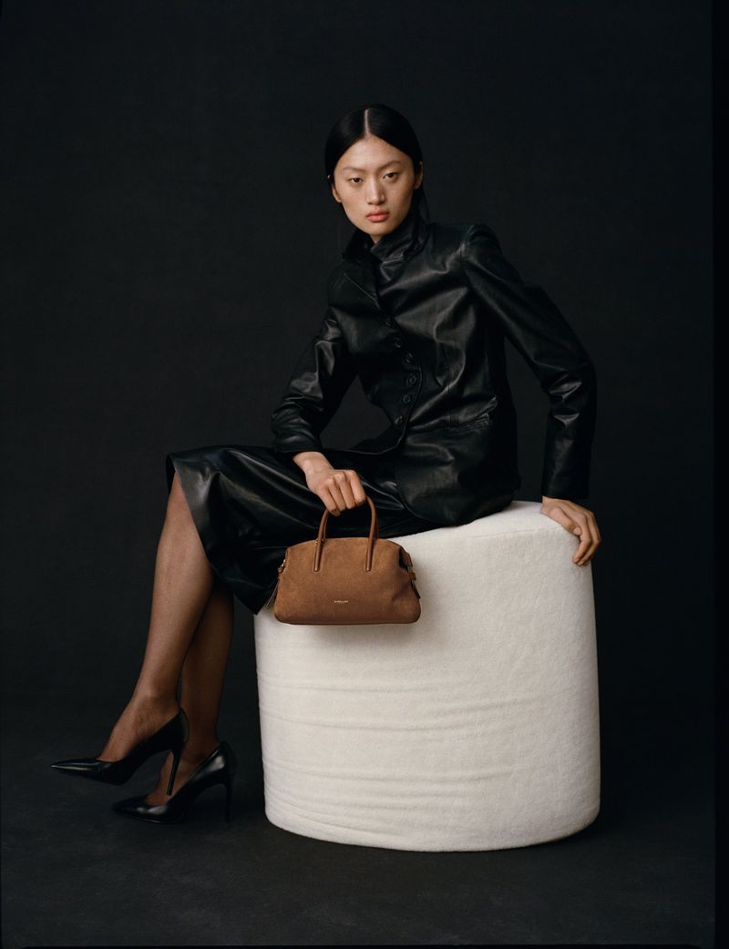 Asian woman in a black leather jacket and skirt sits holding a brown handbag on a white stool against a black background in studio lighting.