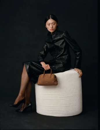 Asian woman in a black leather jacket and skirt sits holding a brown handbag on a white stool against a black background in studio lighting.