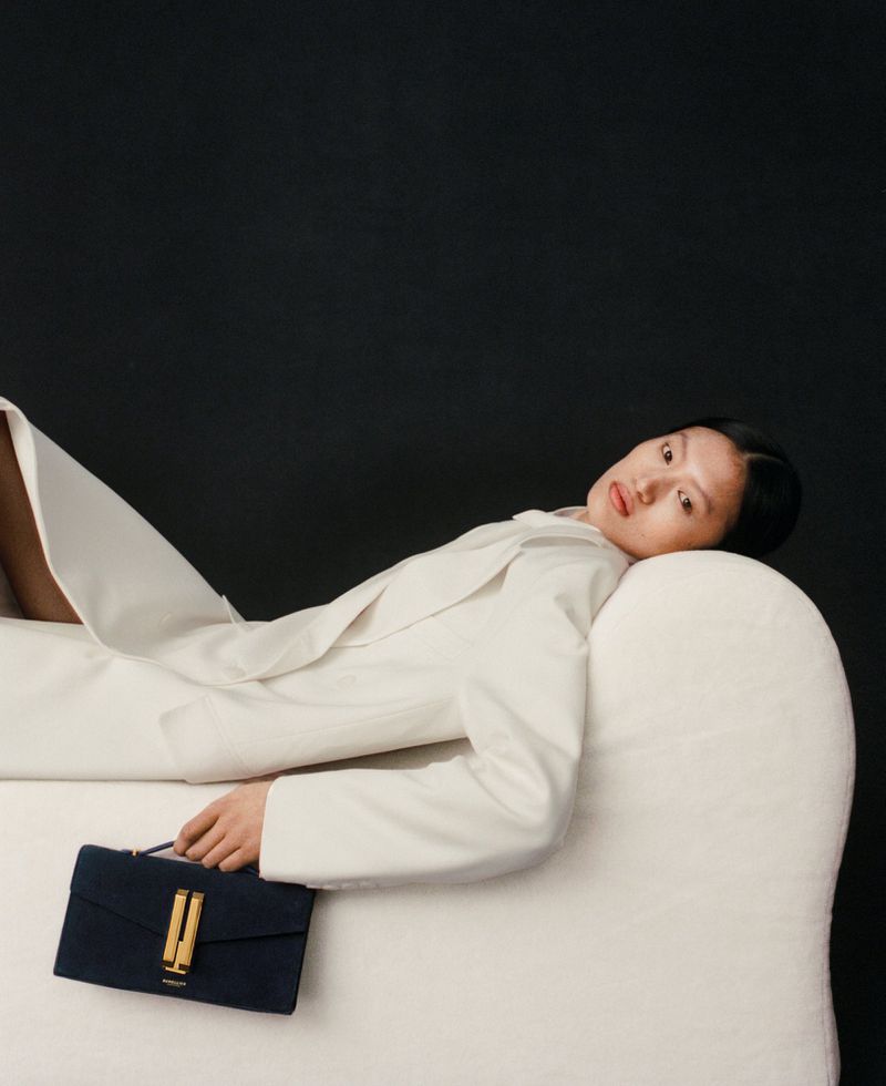 Stylish woman in a white jacket reclines on a sofa with a navy and gold clutch purse against a dark background in a studio setting.