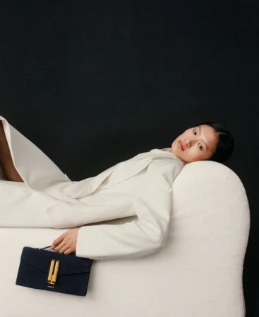 Stylish woman in a white jacket reclines on a sofa with a navy and gold clutch purse against a dark background in a studio setting.