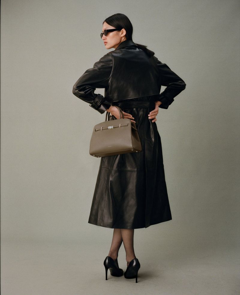 A slender adult woman in a black leather trench coat, sunglasses, and heels carries a structured tan leather handbag in a studio shot.