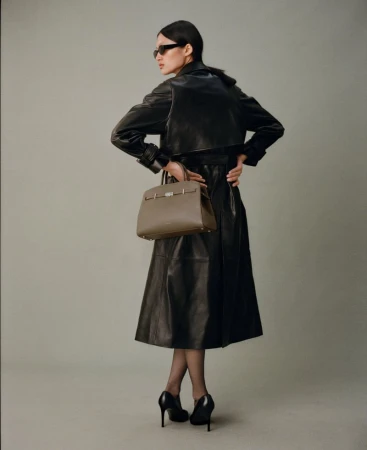 A slender adult woman in a black leather trench coat, sunglasses, and heels carries a structured tan leather handbag in a studio shot.