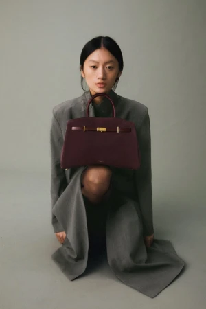 Asian woman kneeling, holding a burgundy leather handbag with gold hardware in a studio setting, wearing a gray oversized suit.