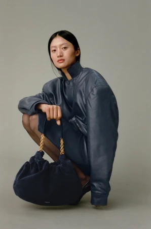 Fashion portrait of an Asian woman crouching with a blue leather jacket and a large navy chain bag on a grey studio backdrop.