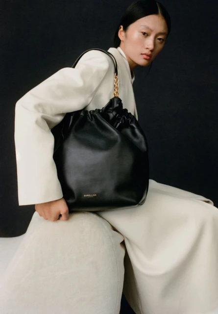 An Asian fashion model presents a black leather bucket bag with a gold chain strap, styled with a cream-colored blazer and wide-leg pants.