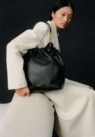 An Asian fashion model presents a black leather bucket bag with a gold chain strap, styled with a cream-colored blazer and wide-leg pants.