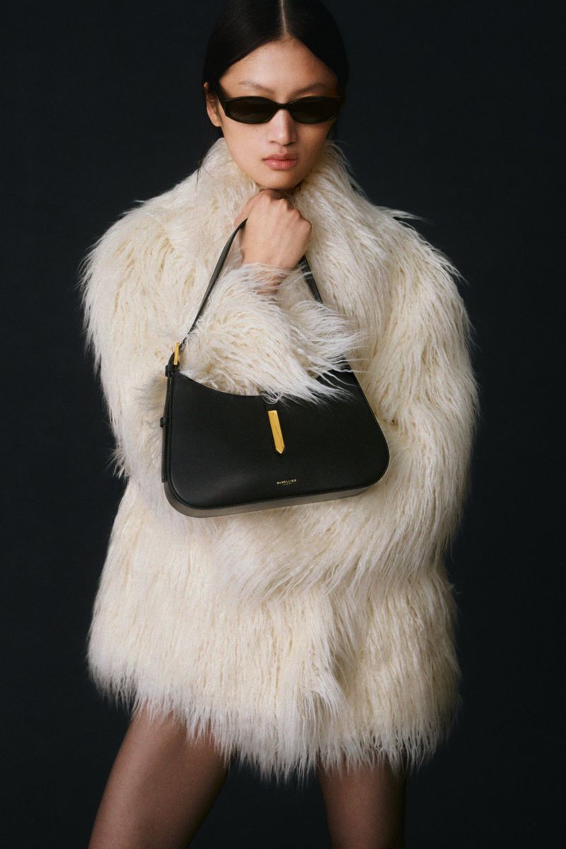 Fashion shot of a woman wearing a fur coat, holding a black leather shoulder bag with gold hardware against a dark backdrop.