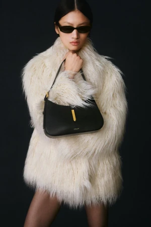 Fashion shot of a woman wearing a fur coat, holding a black leather shoulder bag with gold hardware against a dark backdrop.