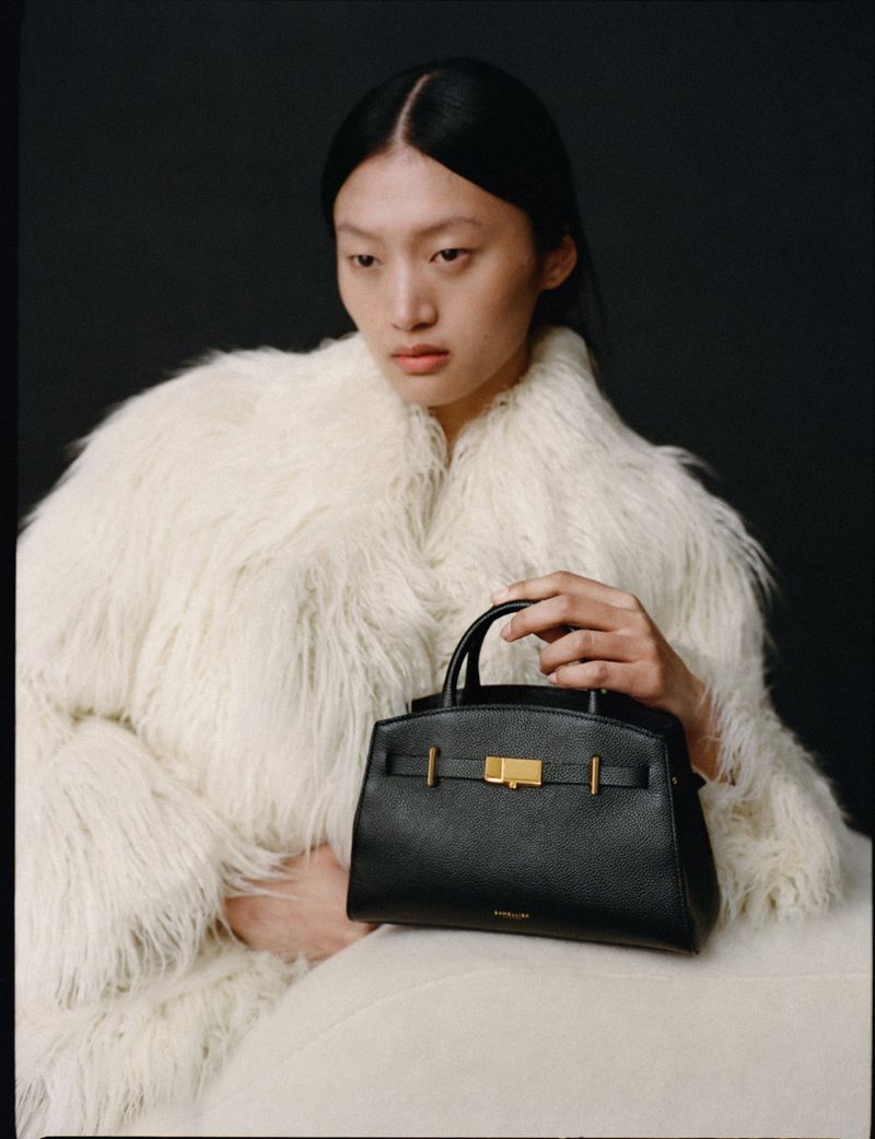 Elegant adult woman in a fur coat poses indoors with a black leather purse with gold hardware against a dark background.