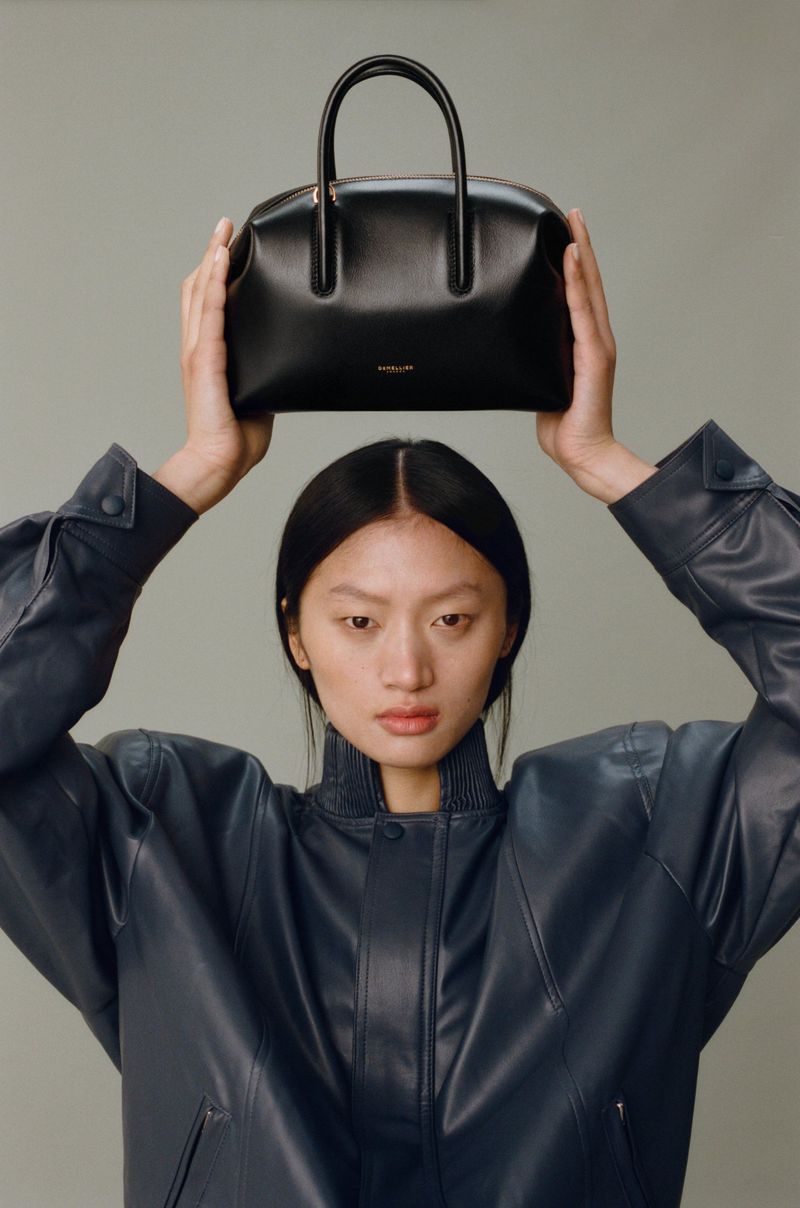 An Asian woman in a navy jacket is holding a black leather purse above her head against a light gray background in a studio shot.