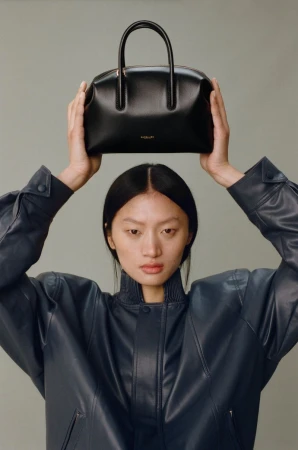 An Asian woman in a navy jacket is holding a black leather purse above her head against a light gray background in a studio shot.