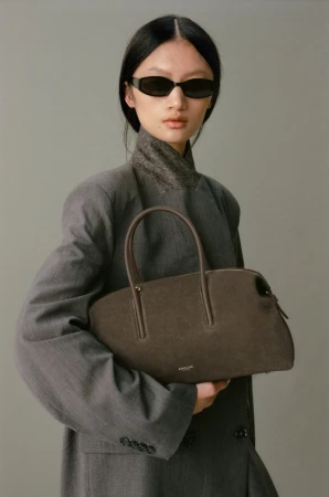 Adult woman in a grey blazer and dark sunglasses holds a brown handbag against a plain grey studio backdrop, fashion editorial.