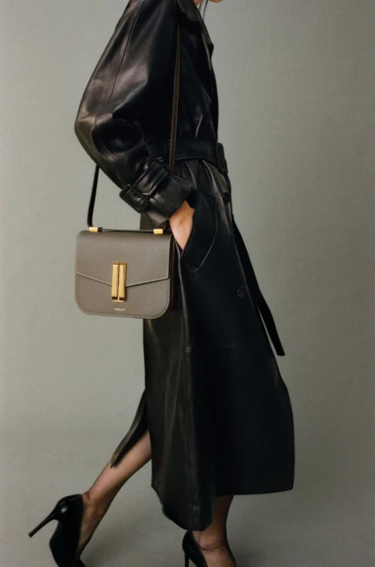 Fashion shot of an adult female in a black leather trench coat, gray leather purse with gold hardware, and black high-heeled shoes.