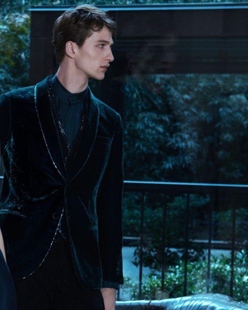 Portrait of a young adult man in a dark velvet jacket, standing indoors with a backdrop of plants visible through a window.