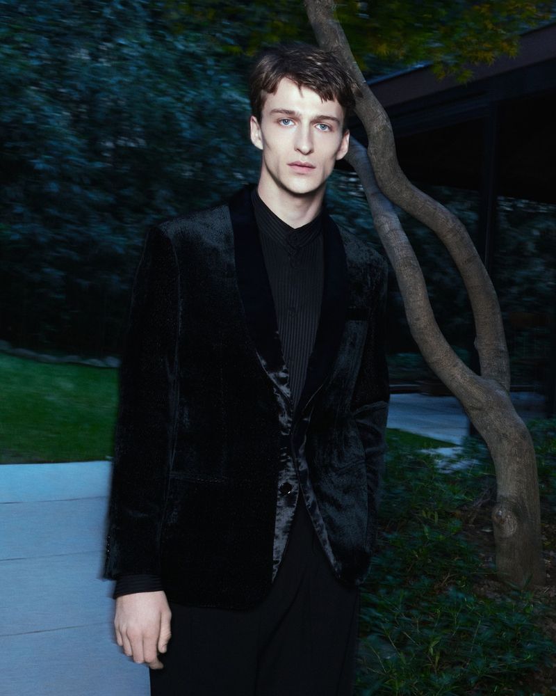 Portrait of a young adult male in a black velvet jacket and shirt, posed outdoors against a backdrop of green foliage and a bare tree.