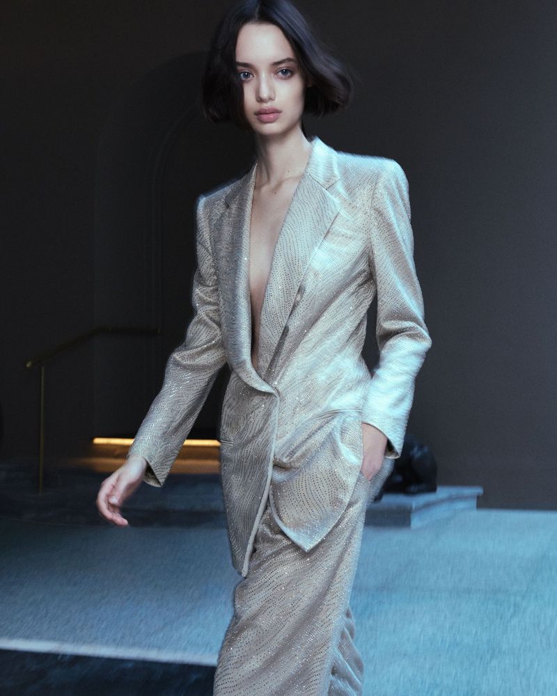 Elegant brunette woman in a sparkly gold pant suit on a grey background with muted lighting, creating a sophisticated fashion image.