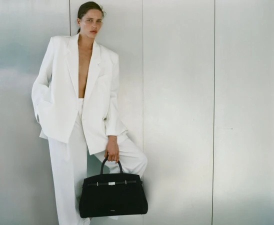 Woman in a white pantsuit poses against a textured wall holding a large black handbag, fashion photography, studio lighting.