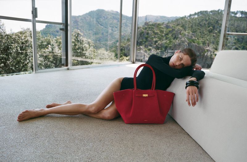 Woman in bodysuit poses next to a red tote bag indoors with a window and mountain view, creating an elegant fashion image.