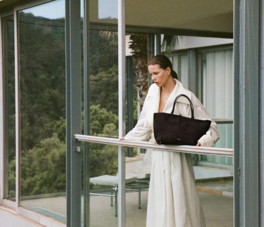 Woman in off-white coat poses on patio holding dark woven tote bag against a green foliage background on a sunny day.
