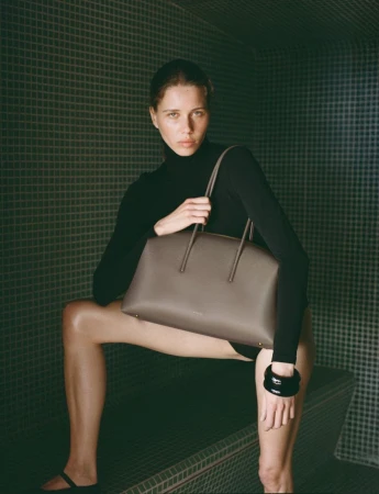 Fashion shot of a young woman in a black turtleneck, holding a grey handbag while seated indoors with a tiled background.
