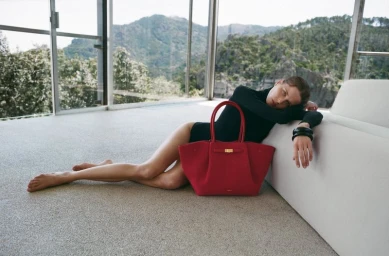 Model wearing a black top and dark bangles reclining barefoot on a textured floor beside a large red handbag and a white sofa.