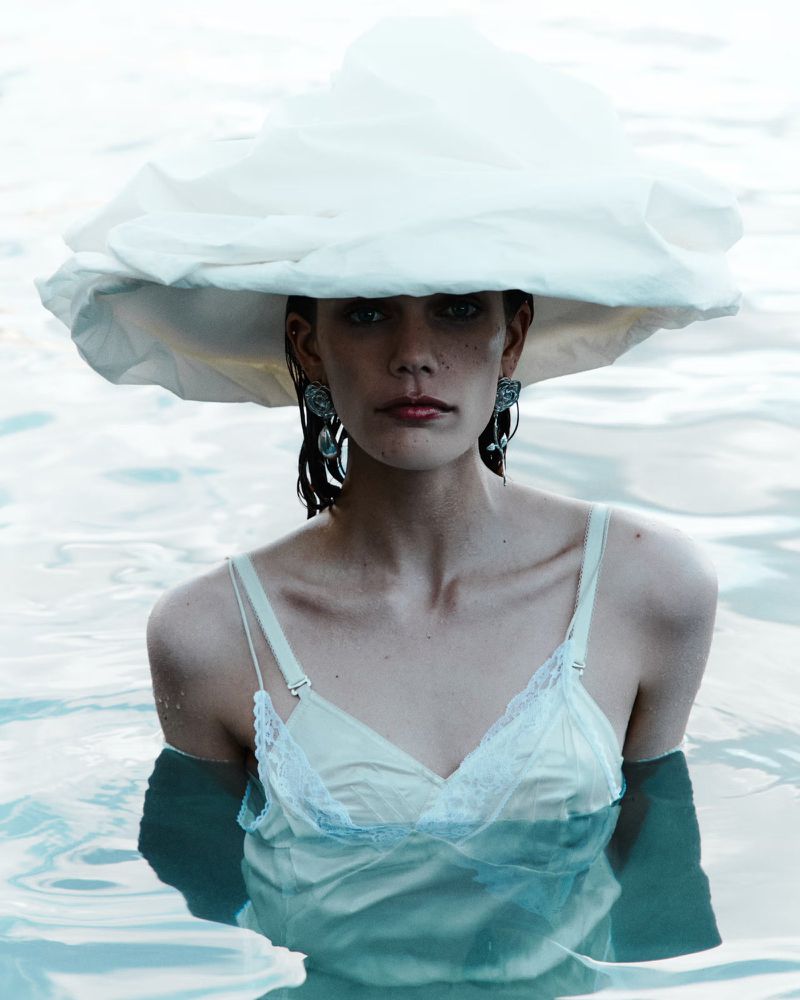A woman in a light-colored slip dress stands in the water, wearing statement earrings and a large white hat.