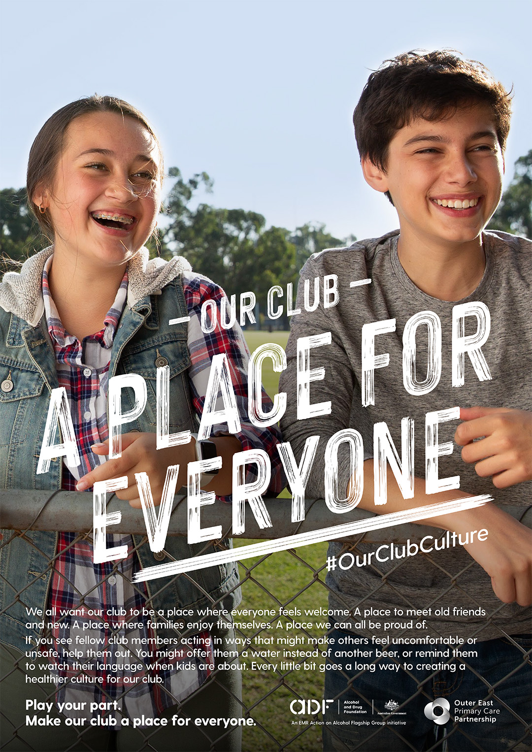 Two smiling teenagers stand outdoors in front of a promotional poster promoting a welcoming club environment for everyone.