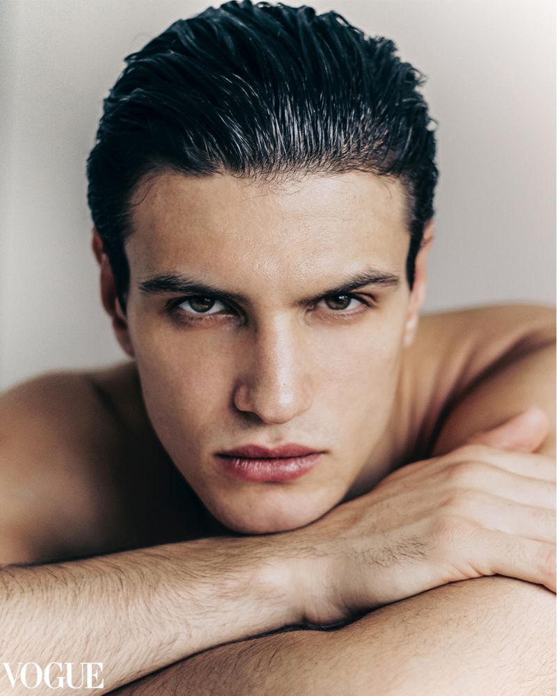 Close-up portrait of a young adult man with wet dark hair slicked back, resting his chin on his crossed arms, with a Vogue logo visible.