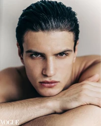 Close-up portrait of a young man with dark wet-styled hair resting his chin on his crossed arms against a neutral background.