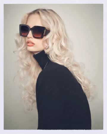 Blonde woman wearing dark oversized sunglasses and a black turtleneck against a neutral studio background