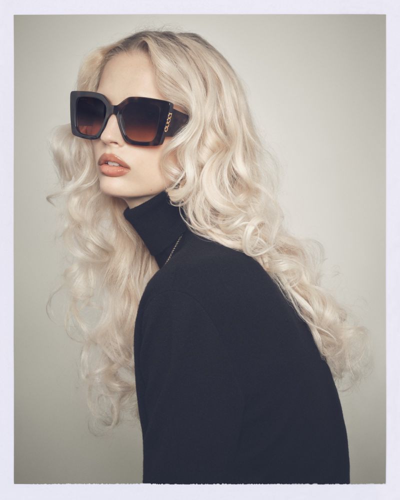 Blonde woman with wavy hair wears oversized square sunglasses and a black turtleneck in a fashion headshot portrait.
