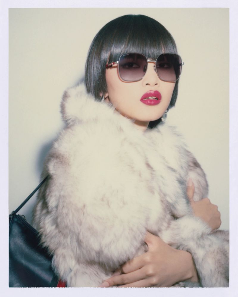 Woman in fur coat and sunglasses against a neutral backdrop, accessorized with earrings and a black bag, with bold red lipstick.