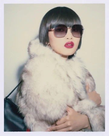 Woman in fur coat and sunglasses against a neutral backdrop, accessorized with earrings and a black bag, with bold red lipstick.