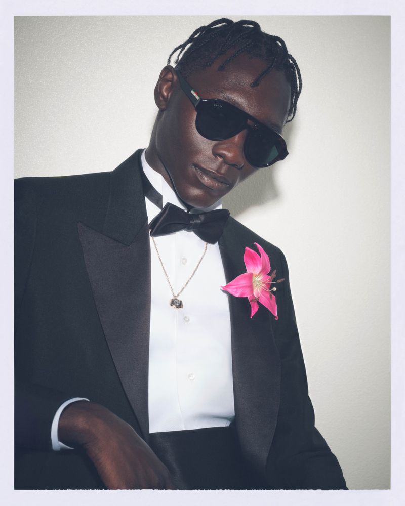 Elegant black man in tuxedo, sunglasses, and floral boutonniere against a plain white background, radiating confident style and sophistication.