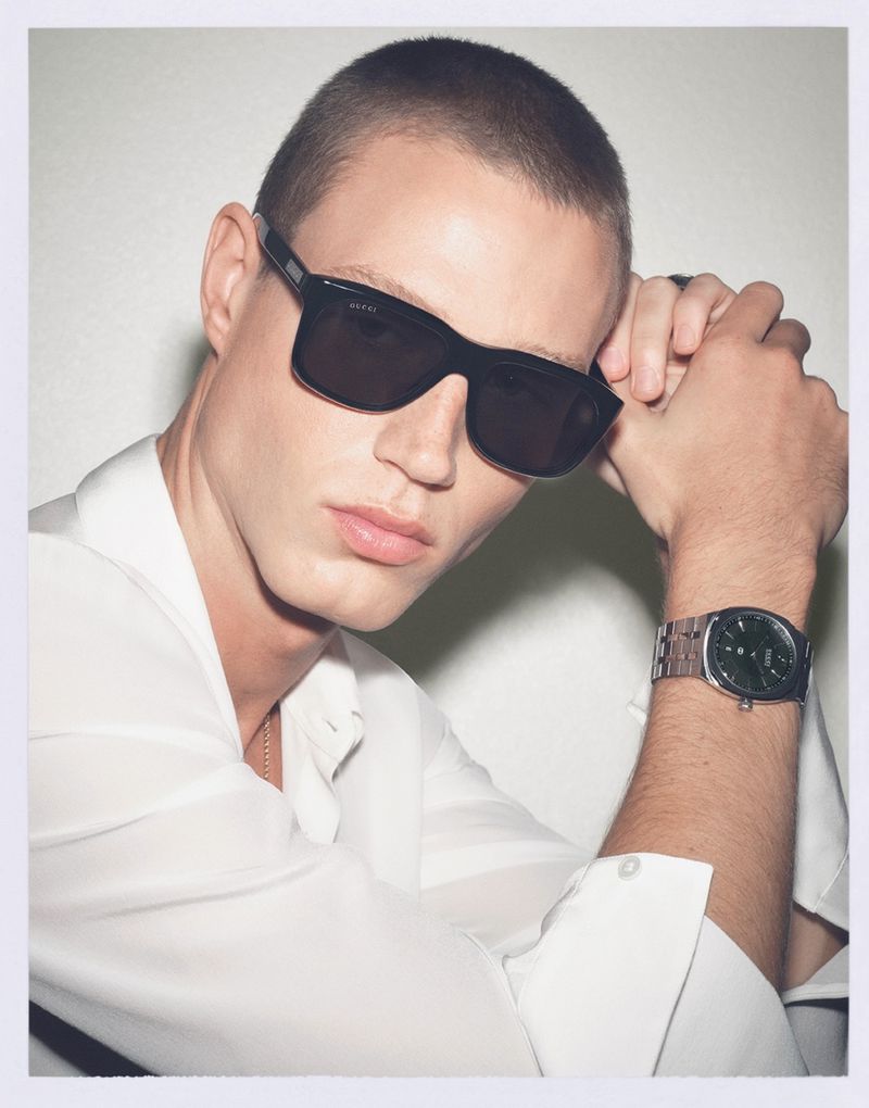 A young adult male model in sunglasses and a white shirt poses indoors, showcasing a wristwatch against a light neutral background.