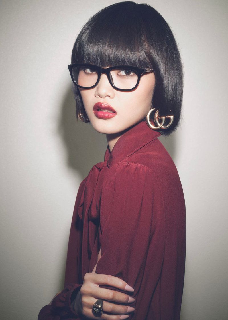 Stylish portrait of a young woman with a dark bob haircut, glasses, and a maroon blouse posing against a neutral backdrop.