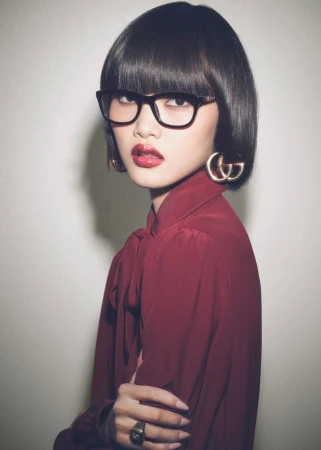 Stylish portrait of a young woman with a dark bob haircut, glasses, and a maroon blouse posing against a neutral backdrop.