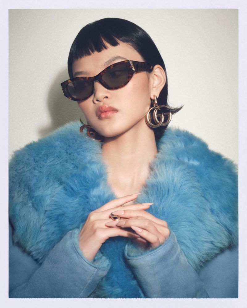 Portrait of an Asian female fashion model wearing sunglasses and a blue fur coat with gold earrings, posing in a studio setting.