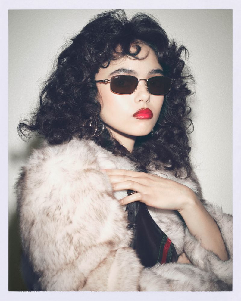 Woman poses in sunglasses and fur coat against a neutral backdrop, showcasing a blend of retro fashion and modern style.