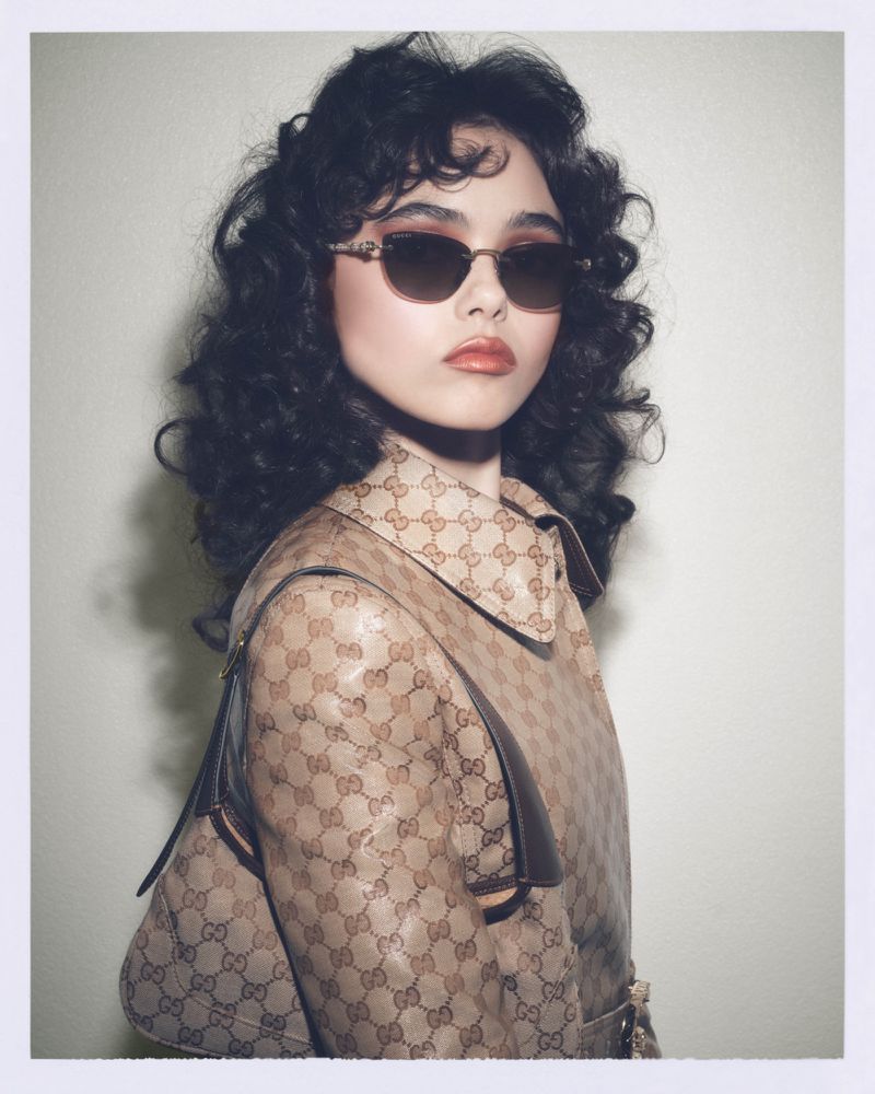 A woman with curly dark hair and sunglasses poses in a Gucci-print coat, accented by a matching purse in a studio setting, fashionably styled.